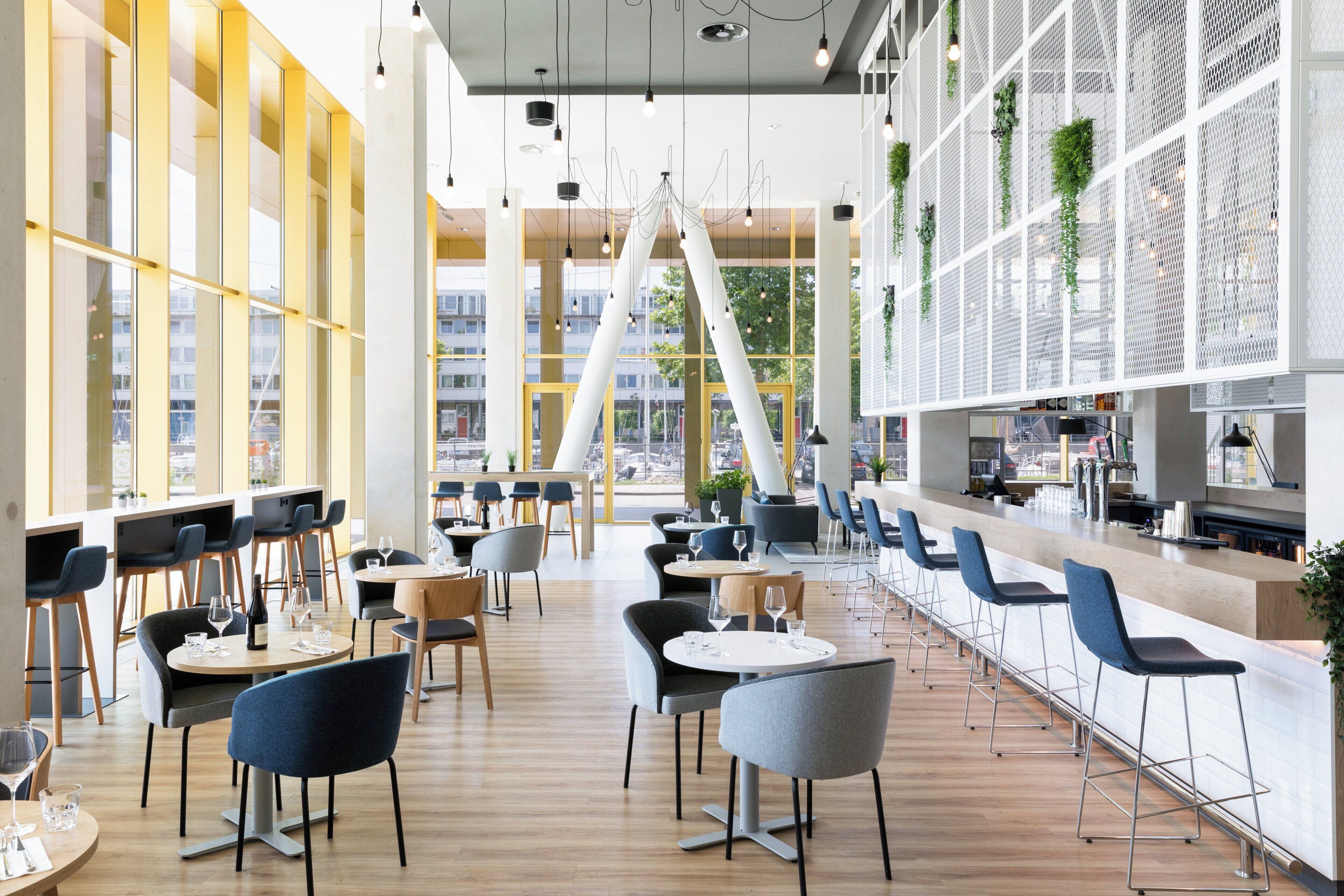 Un restaurante moderno con varias zonas de estar, incluyendo mesas y taburetes de bar, grandes ventanales y vistas a la ciudad.