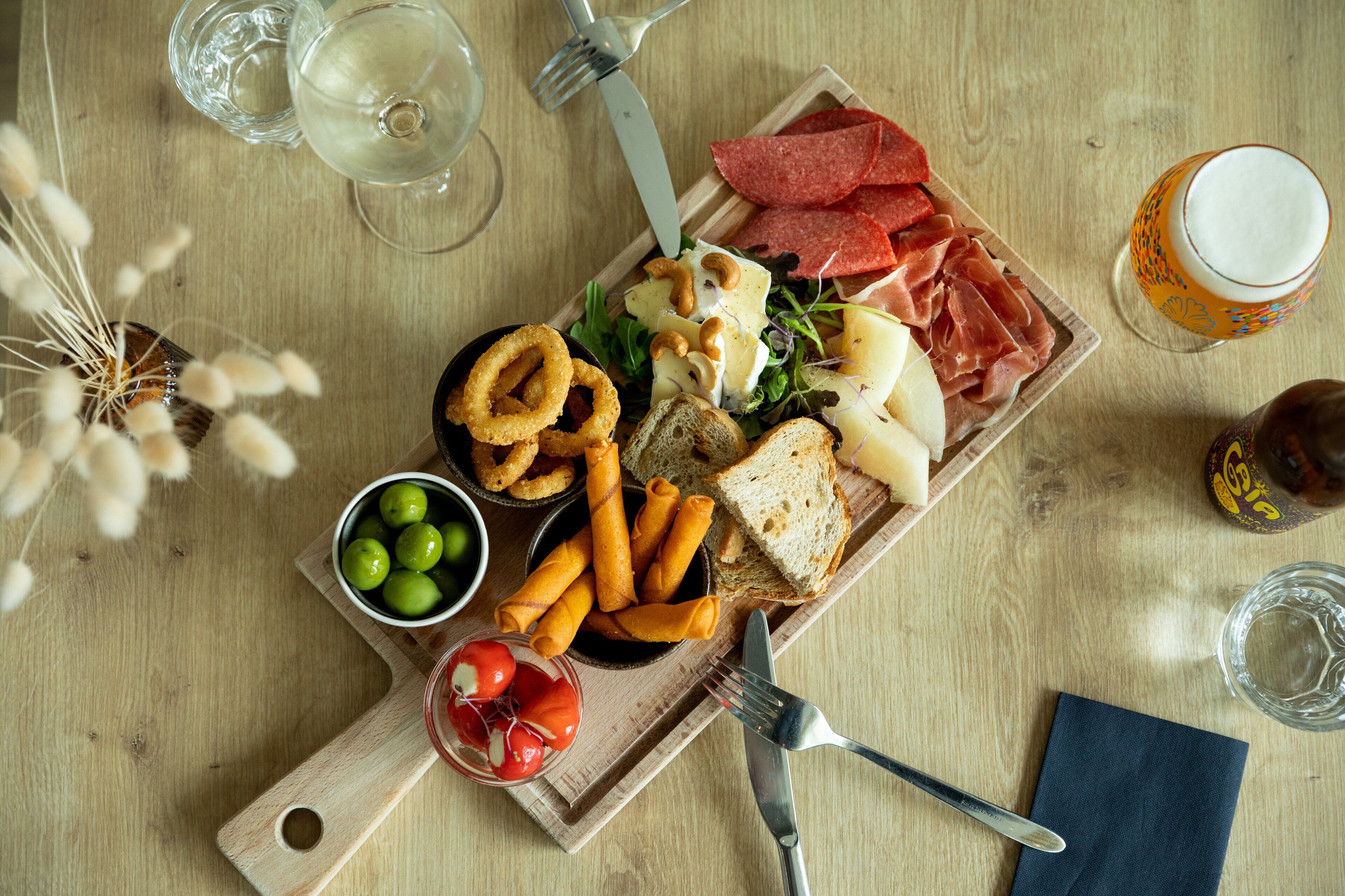 Una mesa de madera con diversos alimentos, como queso, carne y verduras, además de bebidas y utensilios.