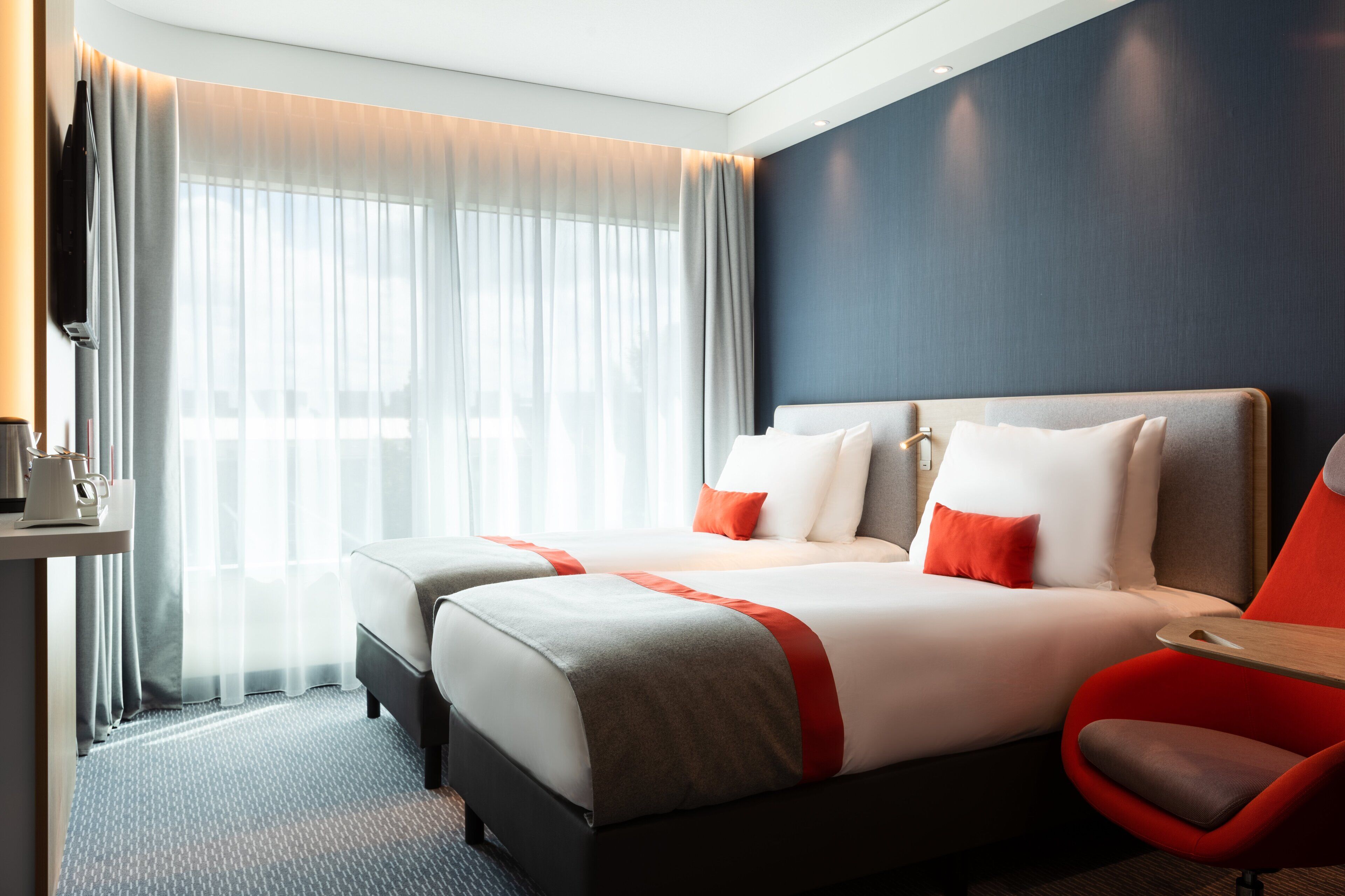 A hotel room with two beds, a TV, a desk with a coffee maker, and a red chair.