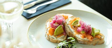 Un plato con dos sándwiches abiertos cubiertos de aguacate, salmón ahumado y verduras encurtidas, acompañado de una copa de vino blanco y cubiertos sobre una mesa.