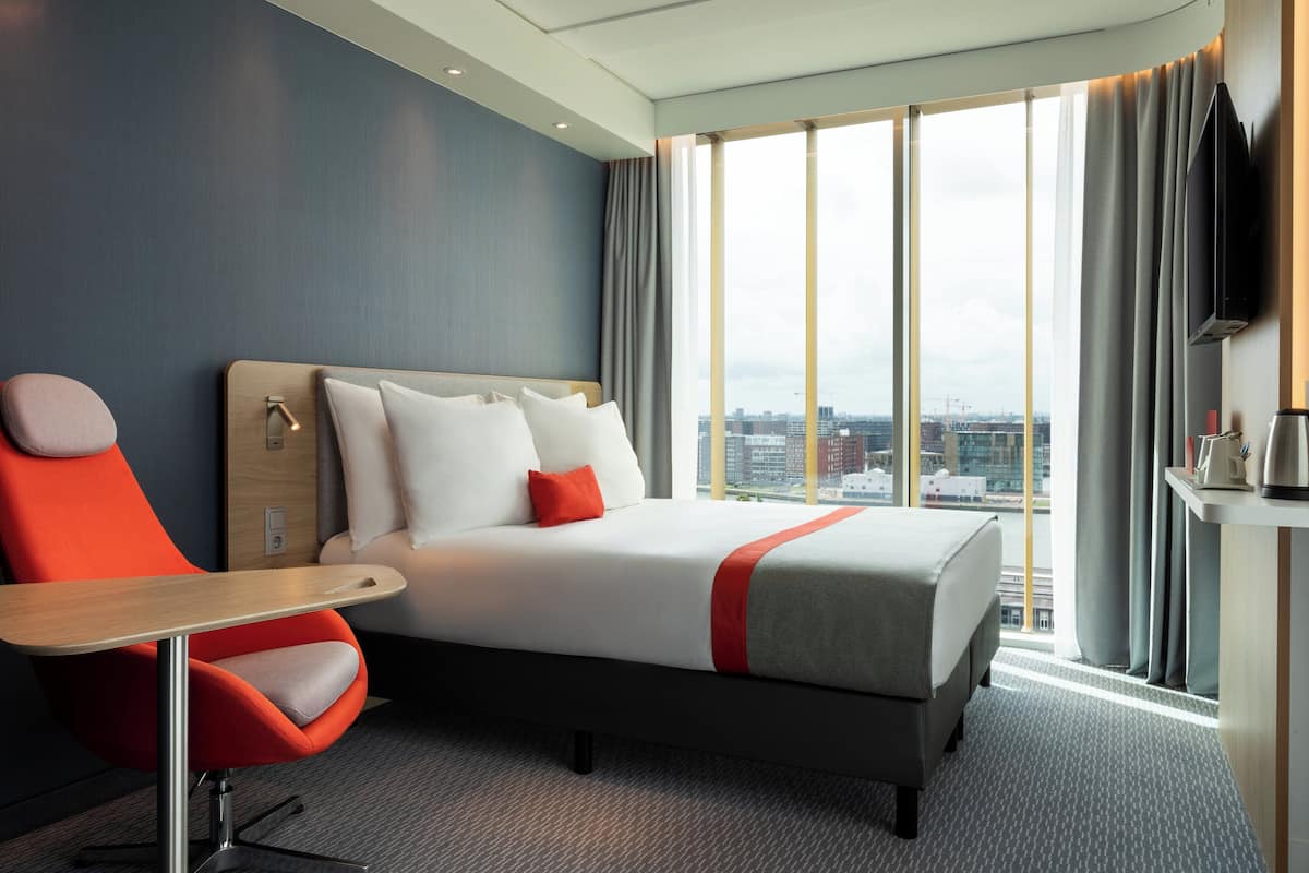 A modern hotel room with a large bed, a desk, a chair, a TV, and a city view.