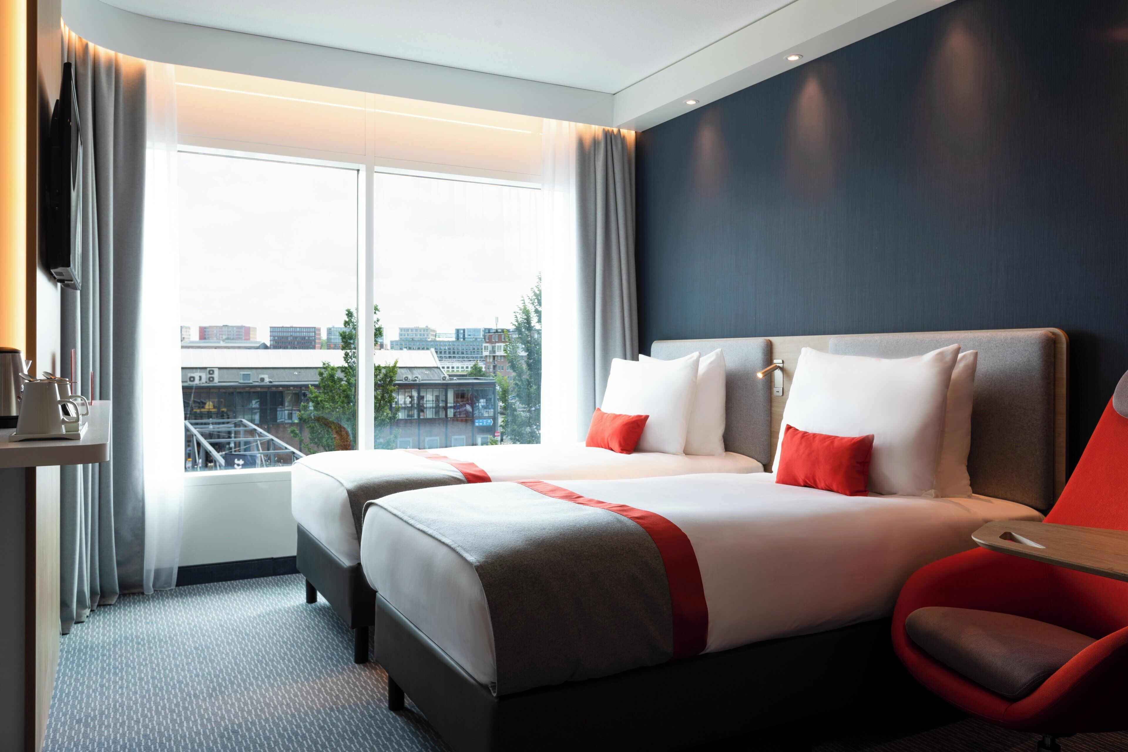 A hotel room with two beds, a red chair, a coffee machine, and a view of buildings through the window.