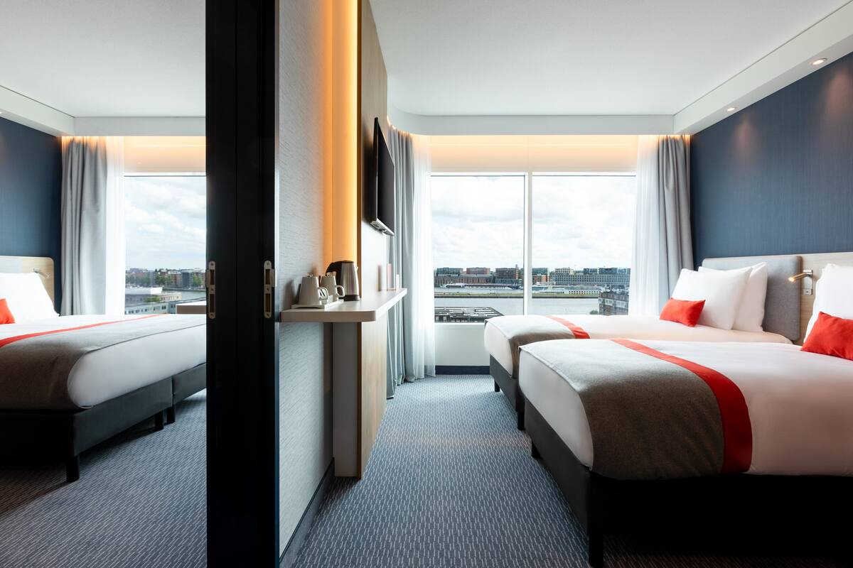 A hotel room with two beds, a desk, and a large window offering a city view.