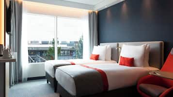 A hotel room with a large window, two beds, a red chair, and a coffee machine.