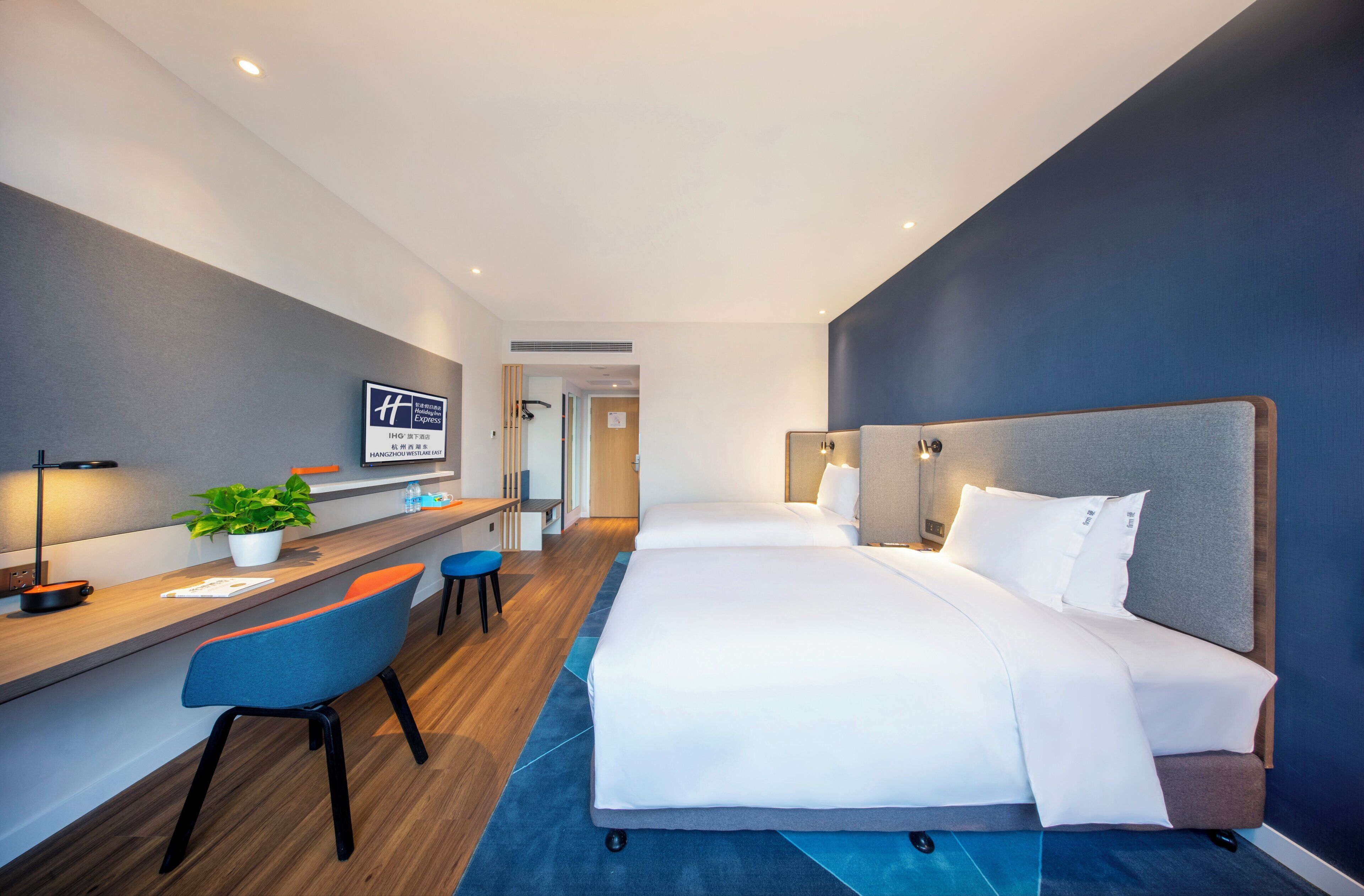 A hotel room with a desk, two beds, a potted plant, and a sign on the wall.
