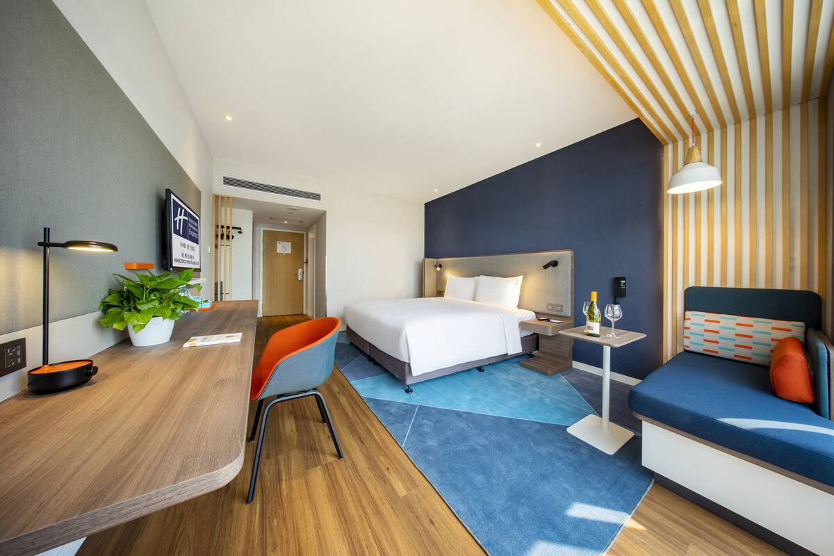 A modern hotel room with a bed, a desk, a chair, a small table with a bottle and glass, and a potted plant.