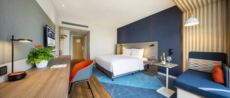 A modern hotel room with a bed, a desk, a chair, a small table with a bottle and glass, and a potted plant.