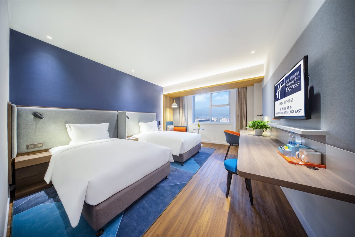 A hotel room with two beds, a desk, a chair, a TV, and a window with a city view.