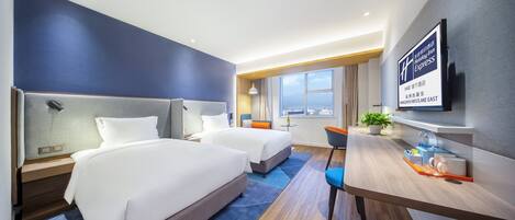 A hotel room with two beds, a desk, a chair, a TV, and a window with a city view.