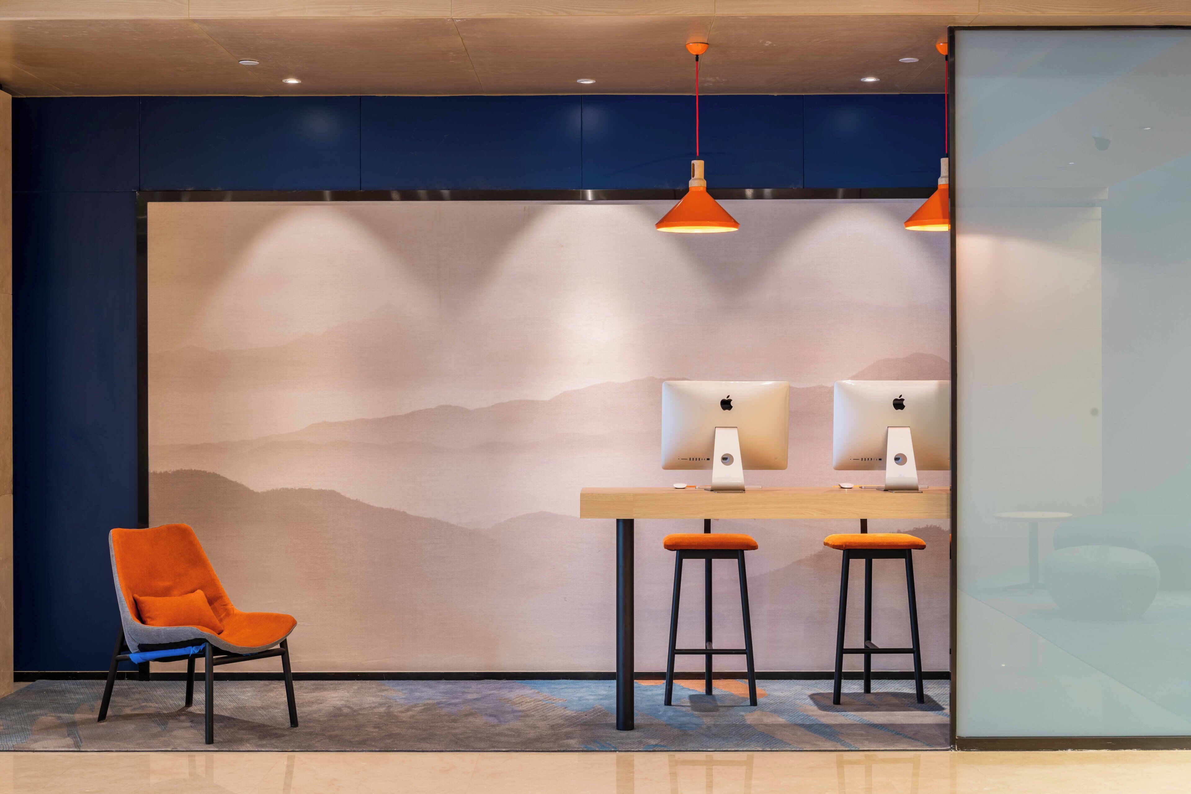 Un espace de travail moderne équipé de deux ordinateurs Apple iMac, d’un bureau en bois, de deux tabourets noirs et d’une chaise orange.