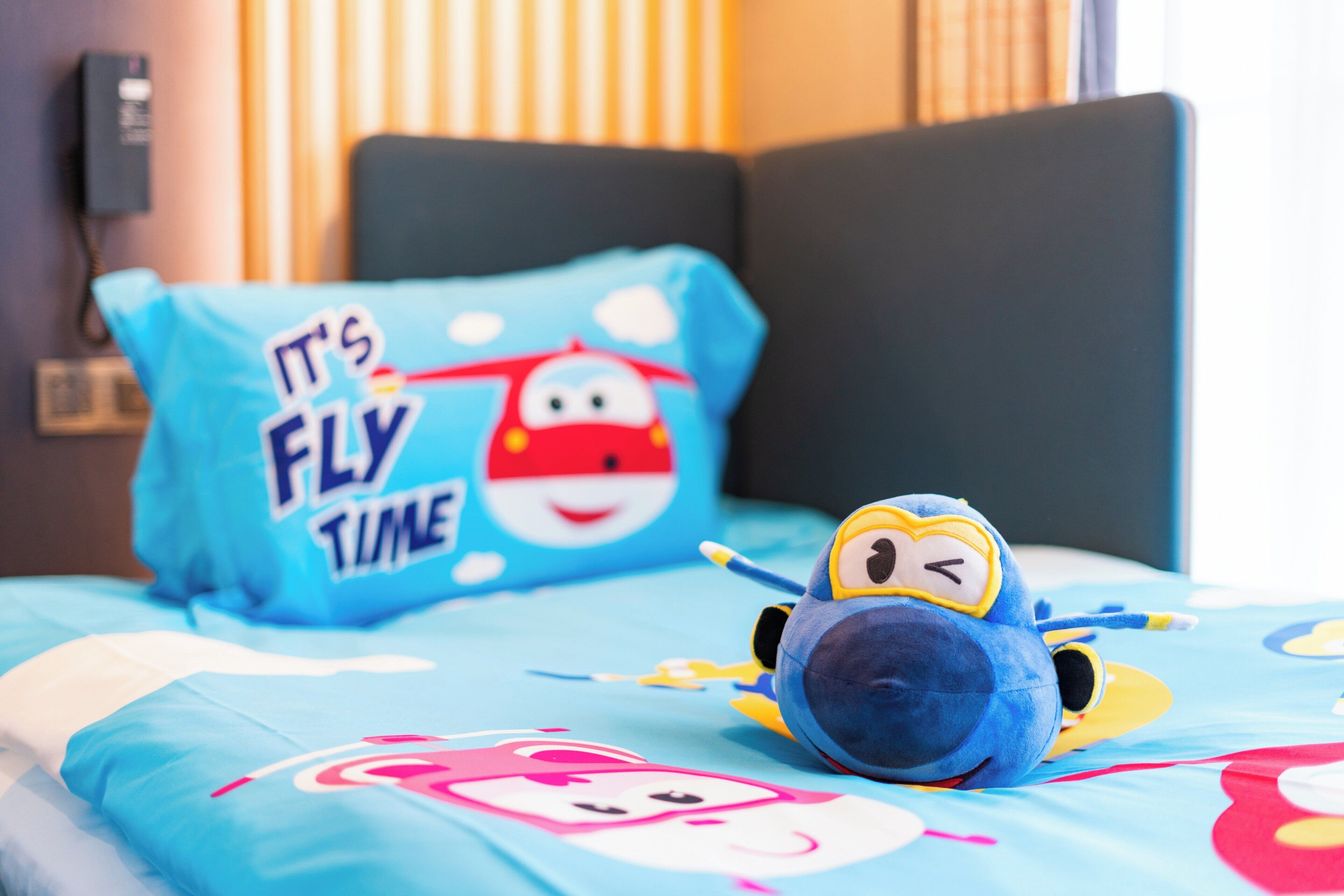 Een bed met een blauw kussen waarop een sneeuwpop afgebeeld is en de tekst 'It's Fly Time', een pluche vogel speelgoed en een dekbedovertrek met tekenfilmfiguren.