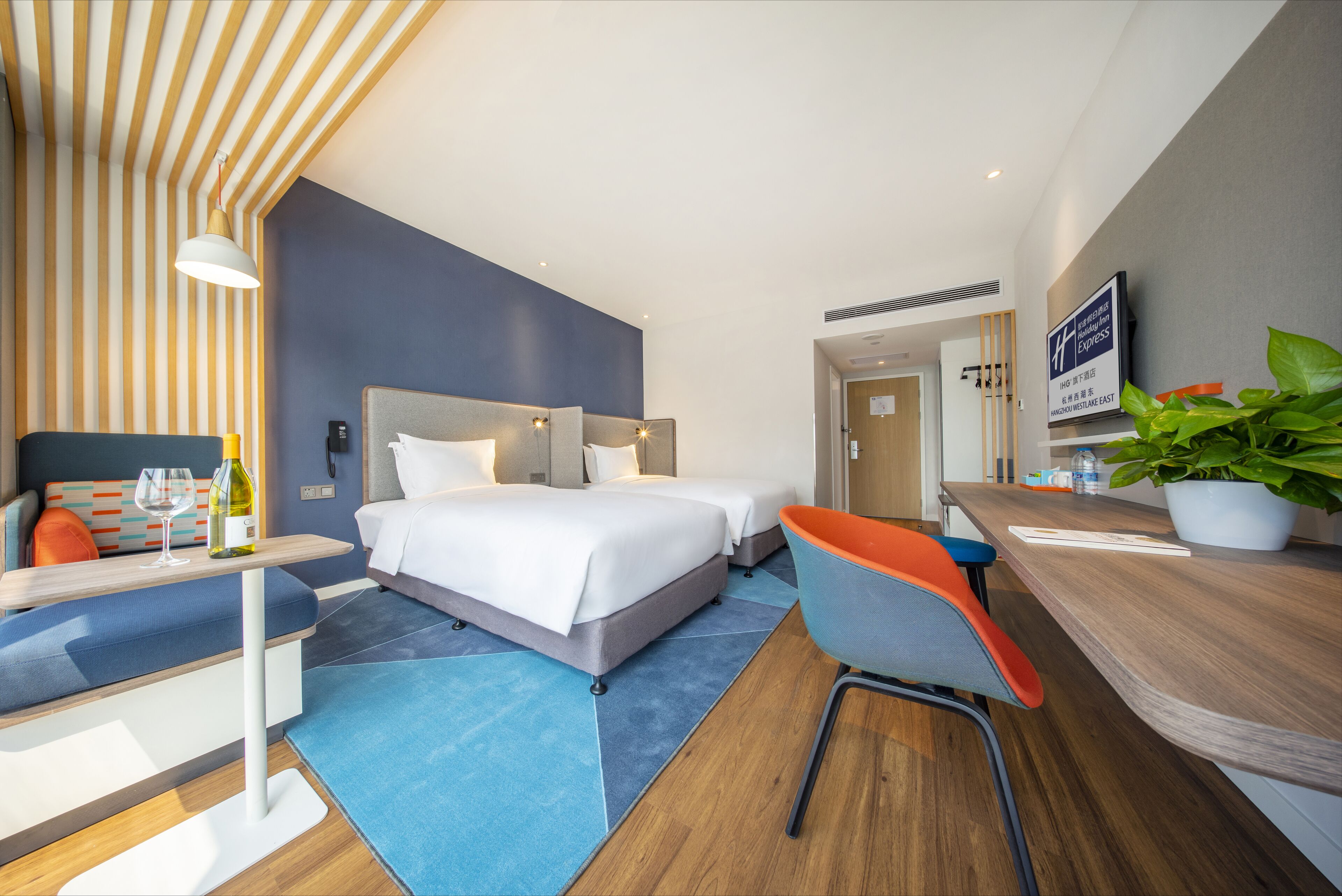 A modern hotel room with a bed, bedside table, chair, and a desk with a plant.