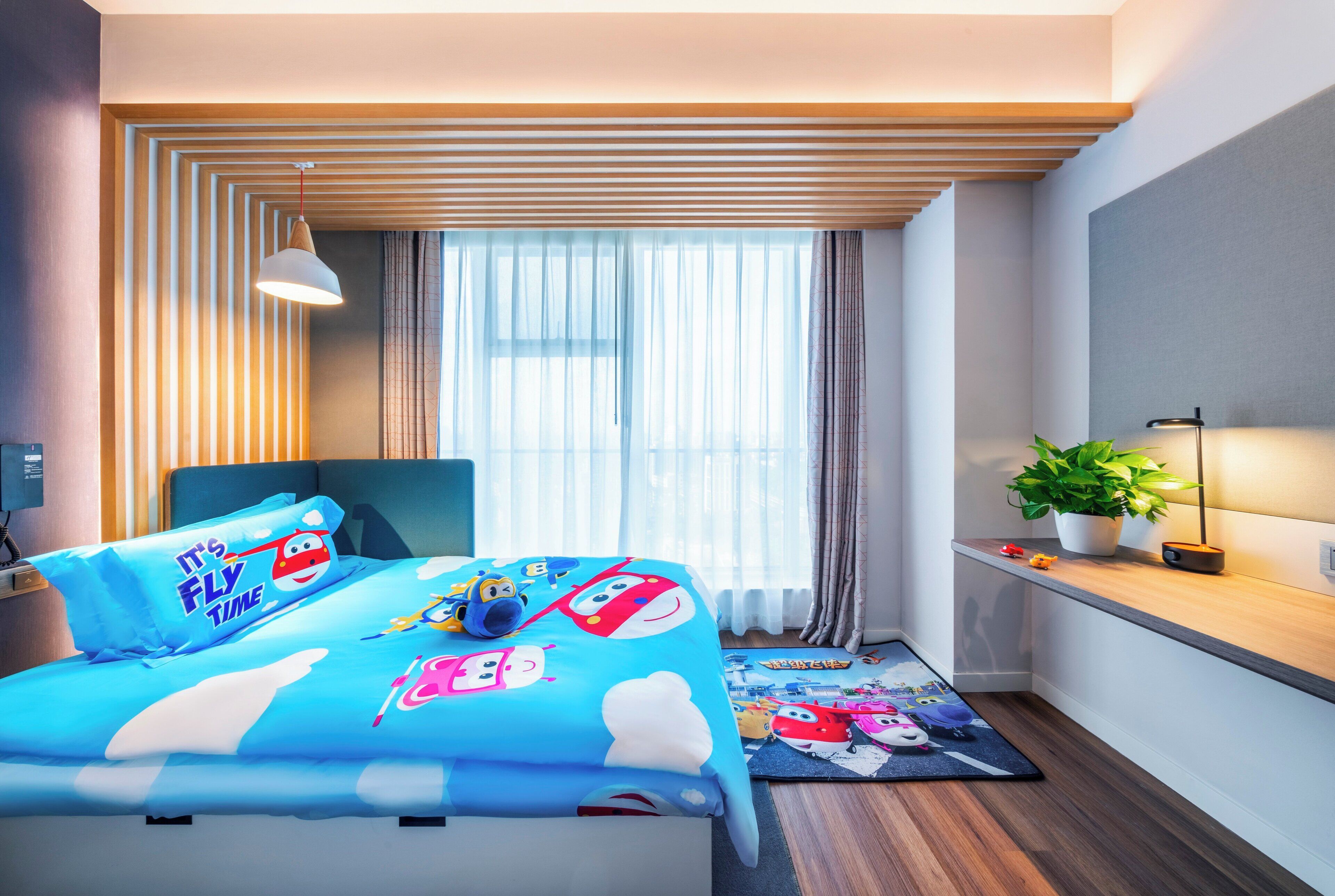 Un dormitorio moderno con cama, mesita de noche, lámpara, ventana con cortinas, planta en maceta y una pequeña estantería con juguetes.