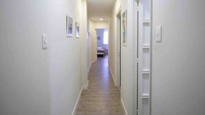 Passadís estret i ben il·luminat amb parets i portes blanques, que condueix a una habitació amb llit i finestra.