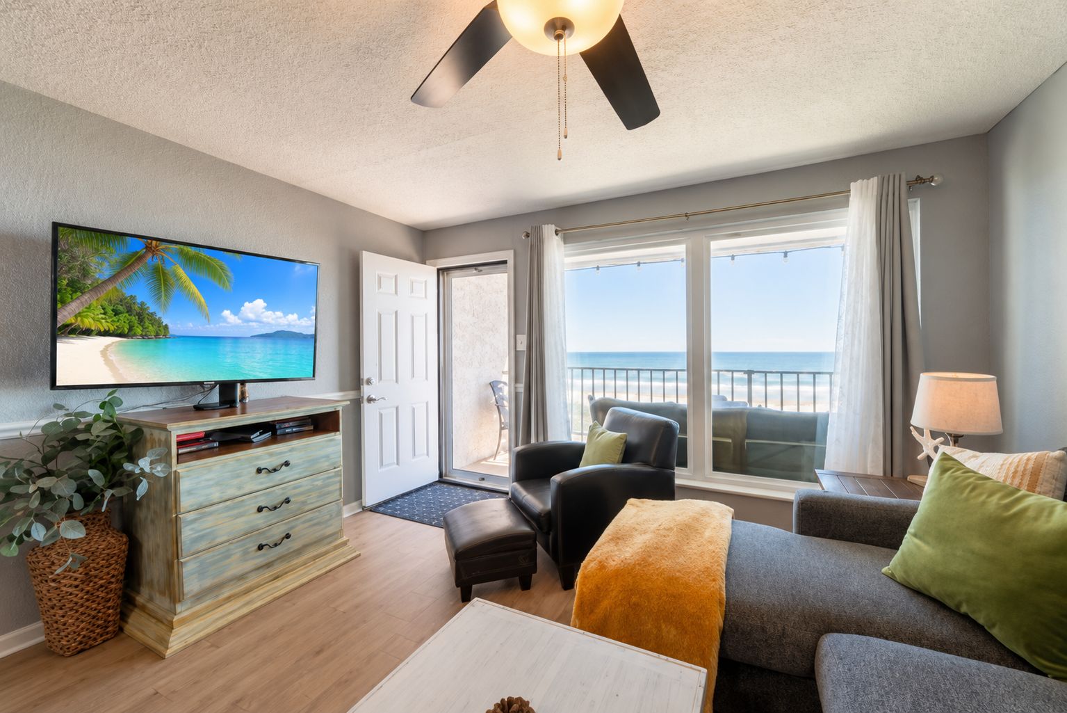 A living room with a television, a sofa, a chair, a coffee table, and a view of the beach through large windows.