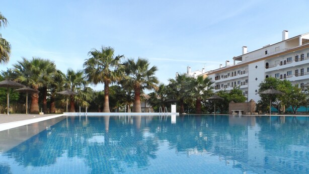 A clear swimming pool with a tiled floor, surrounded by palm trees and umbrellas, with a building in the background. - Turcongel Apartamentos Turisticos (Castro Marim)