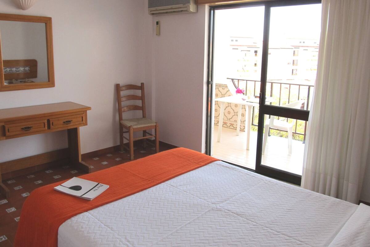A hotel room with a bed, a desk, a chair, and a balcony with a table and chairs.