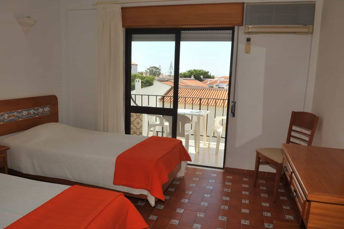 A hotel room with two beds, a dining table, and a sliding glass door leading to a balcony.