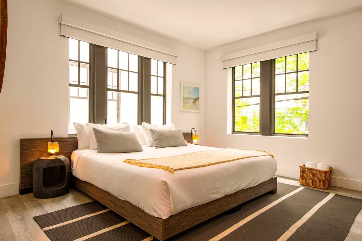 A neatly made bed with a wooden headboard, two bedside lamps, and a large window with a view of greenery.