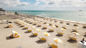 Une plage bordée de rangées de chaises de plage et de parasols jaunes et blancs, une tour de surveillance des sauveteurs et un navire visible au loin.