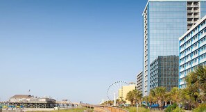 Un paysage urbain côtier avec des immeubles modernes de grande hauteur, une grande roue bien visible et une jetée qui s’avance dans l’eau.