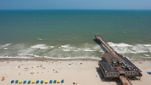 A beach with a pier extending into the ocean, a building with a flat roof, and several beach umbrellas. - Ocean Front resort next to Myrtle Beach Sky Wheel! (Myrtle Beach)