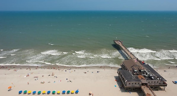 Une plage avec une jetée qui s’avance dans l’océan, un bâtiment doté d’un toit plat et plusieurs parasols de plage.
