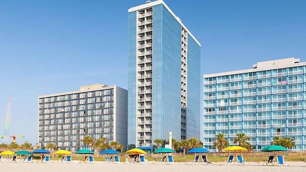A hotel with multiple high-rise buildings, a beach area with sun loungers and umbrellas, and a clear blue sky. - Ocean Front resort next to Myrtle Beach Sky Wheel! (Myrtle Beach)