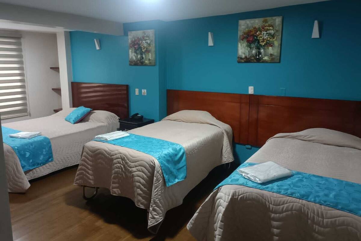 A hotel room with two beds, each with a blue bedspread and white pillows. There are two paintings on the walls, a window with blinds, and a wooden floor.