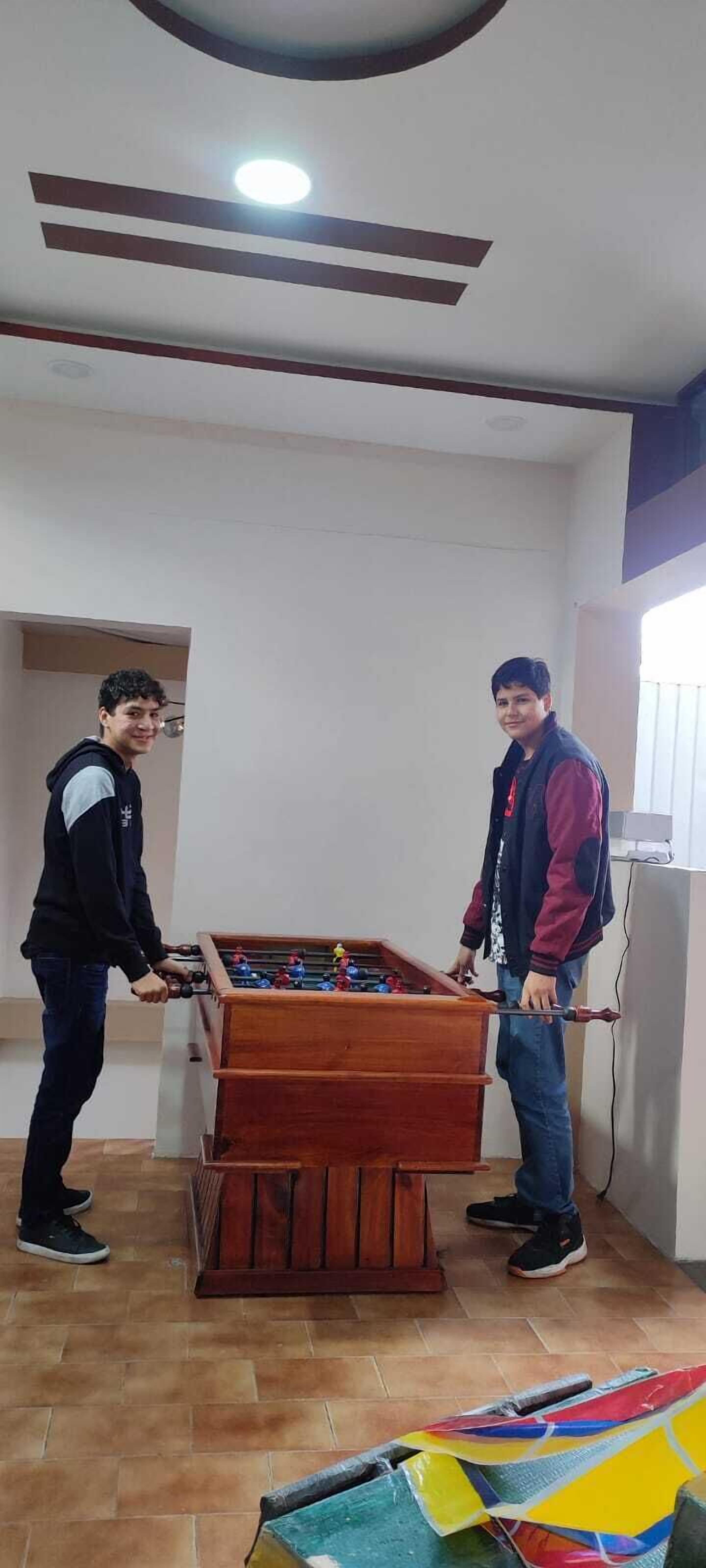 Dos personas jugando al fútbol de mesa en una habitación con suelo de madera, una lámpara en el techo y una ventana.