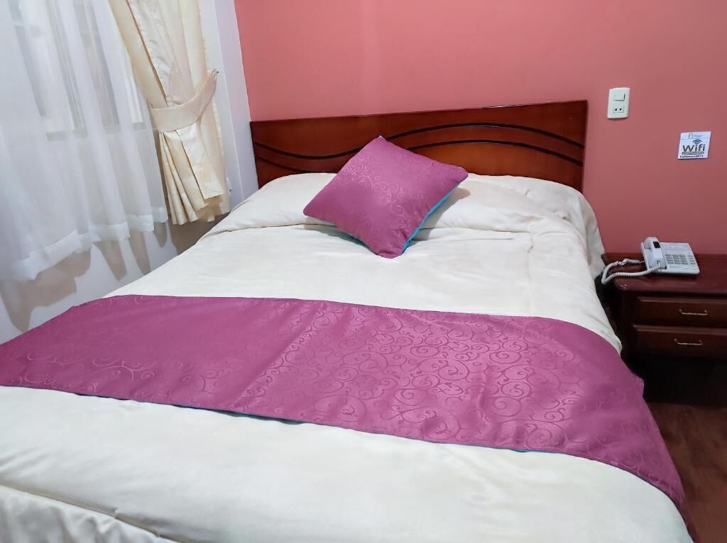 A neatly made bed with a pink and white bedspread, a matching pillow, and a wooden headboard. A bedside table with a phone and a sign indicating WiFi availability is visible.