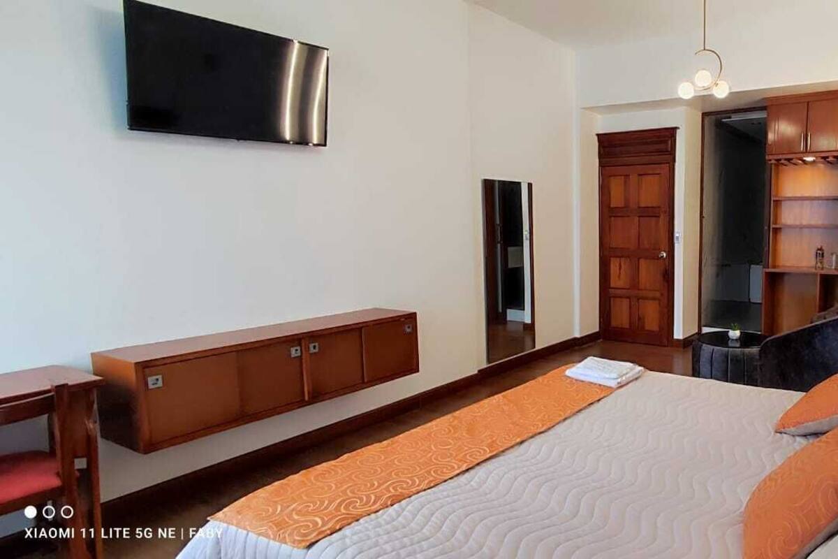 A modern bedroom with a bed, a wooden wardrobe, a TV mounted on the wall, and a small table.