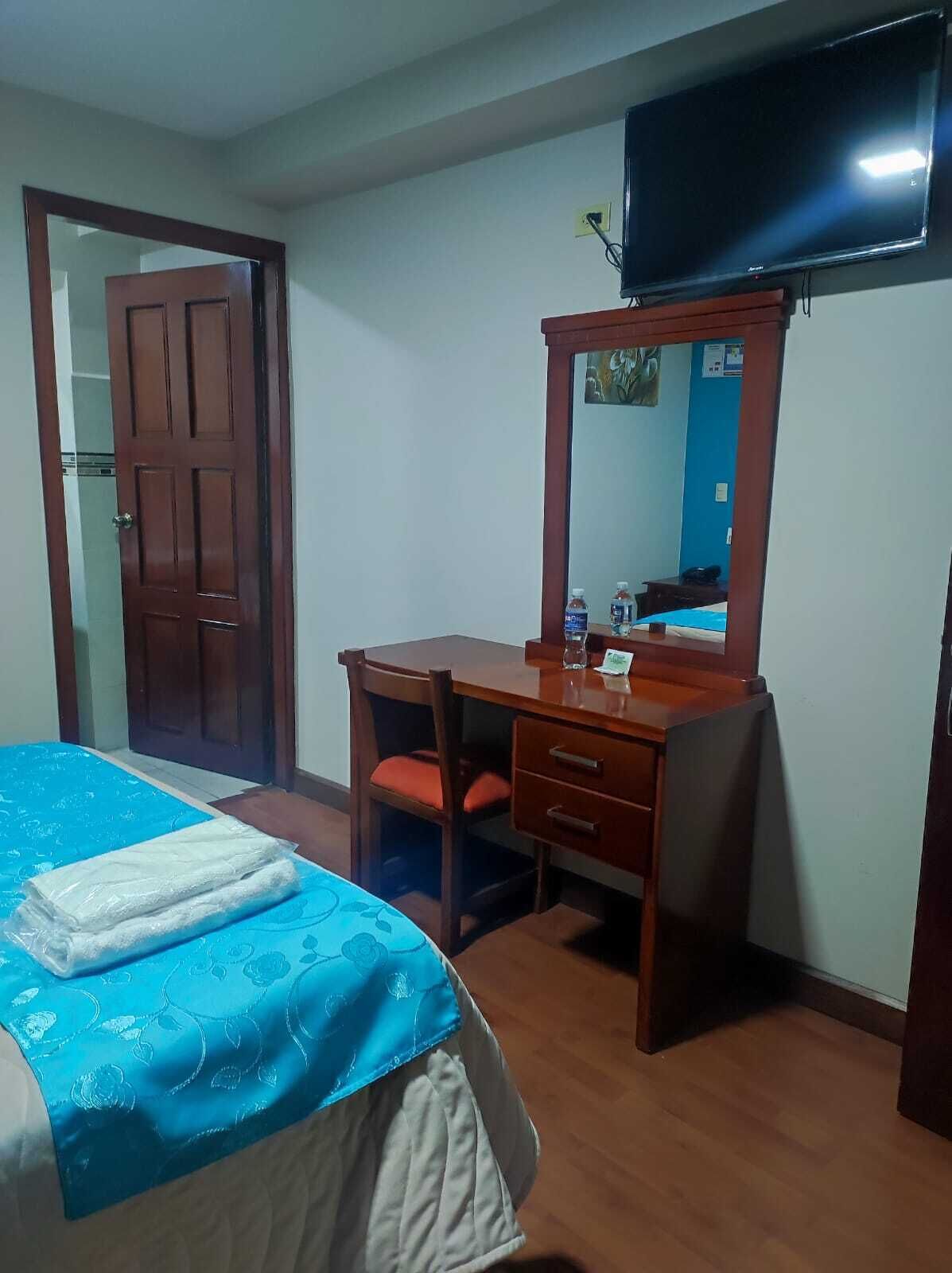 A hotel room with a bed, a wooden desk, a chair, a mirror, and a television on the wall.