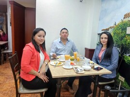 Tres personas sentadas en una mesa con alimentos para el desayuno, incluyendo platos, tazas y un vaso de jugo de naranja.