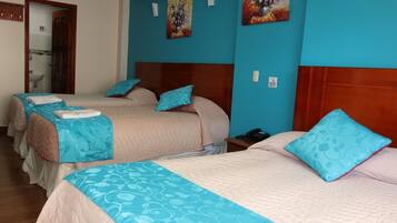 Habitación de hotel con tres camas, cada una con una colcha azul y blanca, y cabeceros de madera. Hay dos cuadros en la pared, una puerta a la izquierda y una mesita de noche con un teléfono.