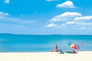 A beach with clear blue water, a few people, and a colorful umbrella on the sand.