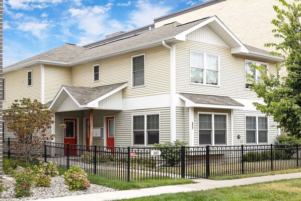 A two-story residential building with a front porch, a black metal fence, and a small garden area.
