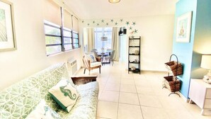 A bright living room with a patterned sofa, a dining area with chairs, a bookshelf, and a wall-mounted shelf. - Seahorse Beach + Ocean Park + Parking (San Juan)