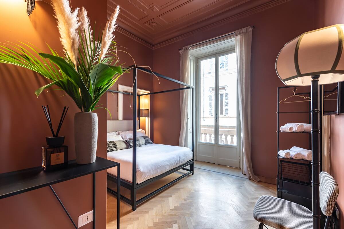 A room with a four-poster bed, a large potted plant, a bedside table with a lamp, and a view of the city through a large window.