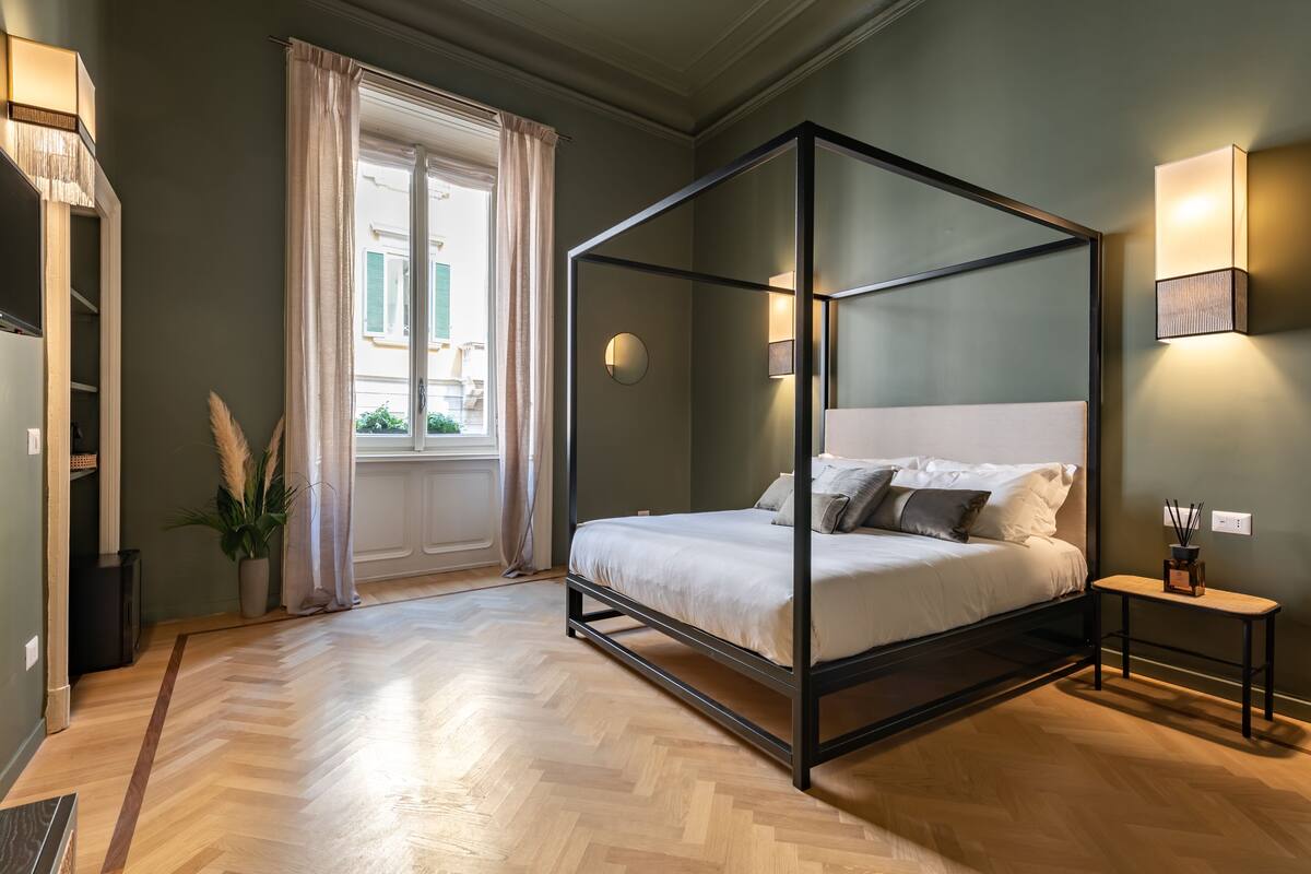 A four-poster bed in a room with a herringbone floor, a window with curtains, and a bedside table with a lamp.