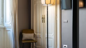 Quarto com porta branca, luminária de chão, cadeira de madeira pequena e TV de tela plana fixada na parede.