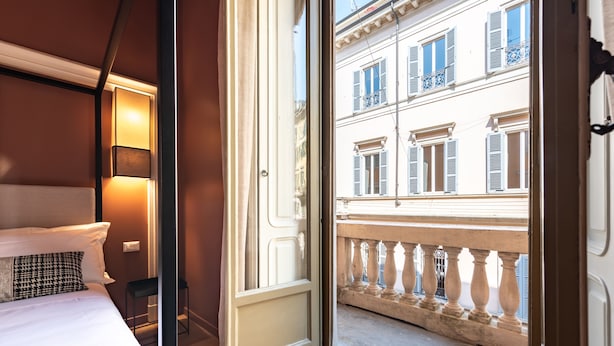 Quarto de hotel com cama, luminária de cabeceira e vista para uma varanda com corrimão e edifícios ao fundo.
