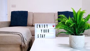 A living room with a beige sofa, a blue throw pillow, a white lightbox with "ENJOY YOUR STAY" text, and a potted fern. - Ye Olde Windmill Apartment Free Parking (York)