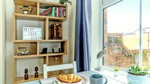 A cozy reading nook with a bookshelf, a table with a plate of croissants, a cup, and a window with a view of a brick wall and greenery. - Ye Olde Windmill Apartment Free Parking (York)