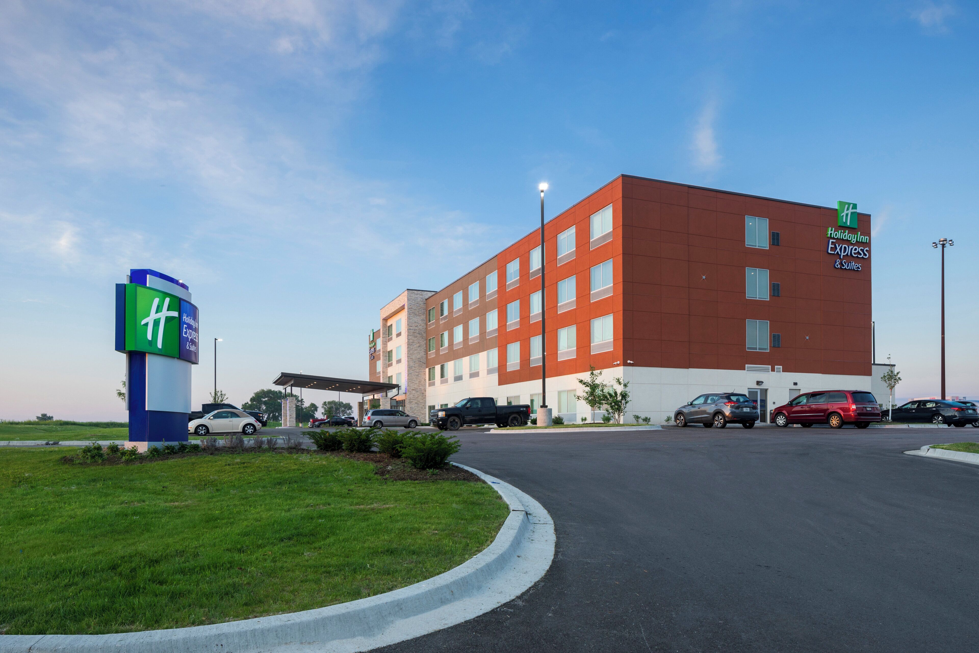 Modernilla punavalkoisella julkisivulla varustettu Holiday Inn Express & Suites -hotelli, jossa on useita parkkipaikkoja ja kirkas taivas.