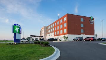 A Holiday Inn Express & Suites hotel with a modern red and white facade, a parking lot with several vehicles, and a clear sky.