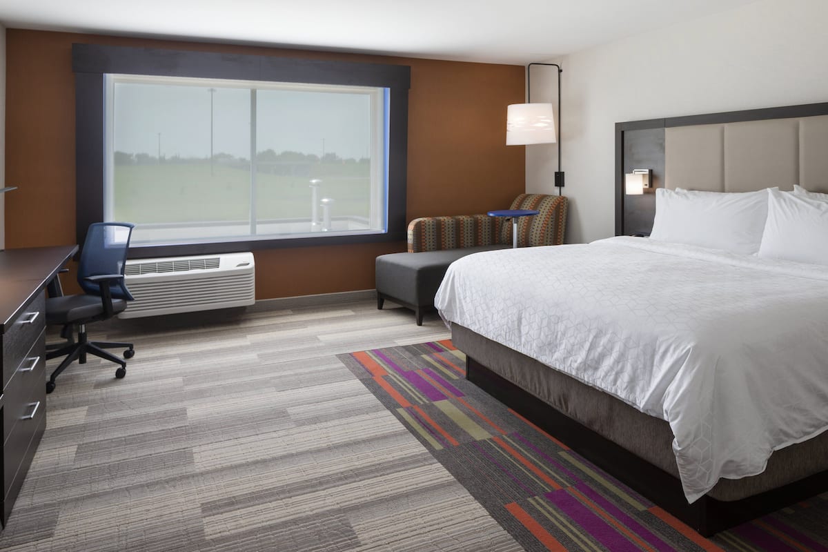 Ein Hotelzimmer mit einem großen Bett, einem Schreibtisch mit Stuhl, einem kleinen Tisch und einem Fenster mit Blick auf ein Feld.