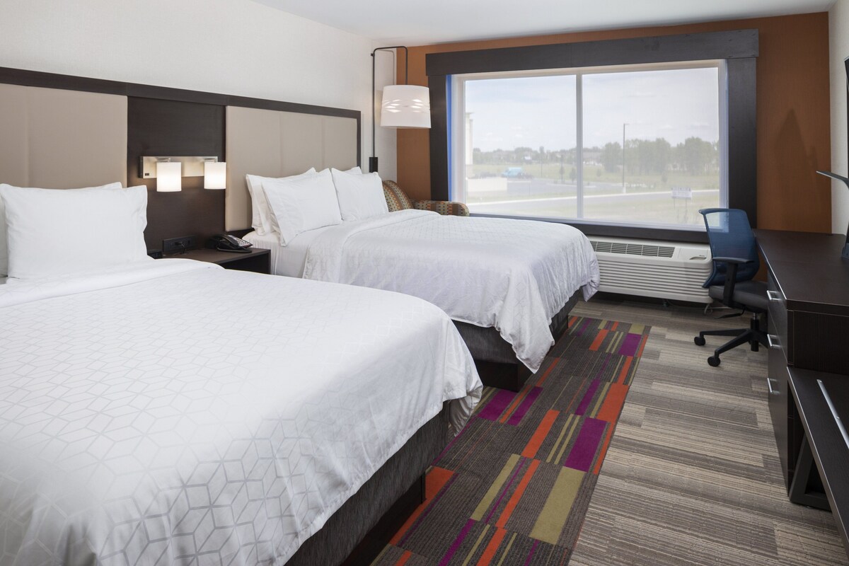 A hotel room with two beds, a desk, a chair, and a large window with a view of the outside.