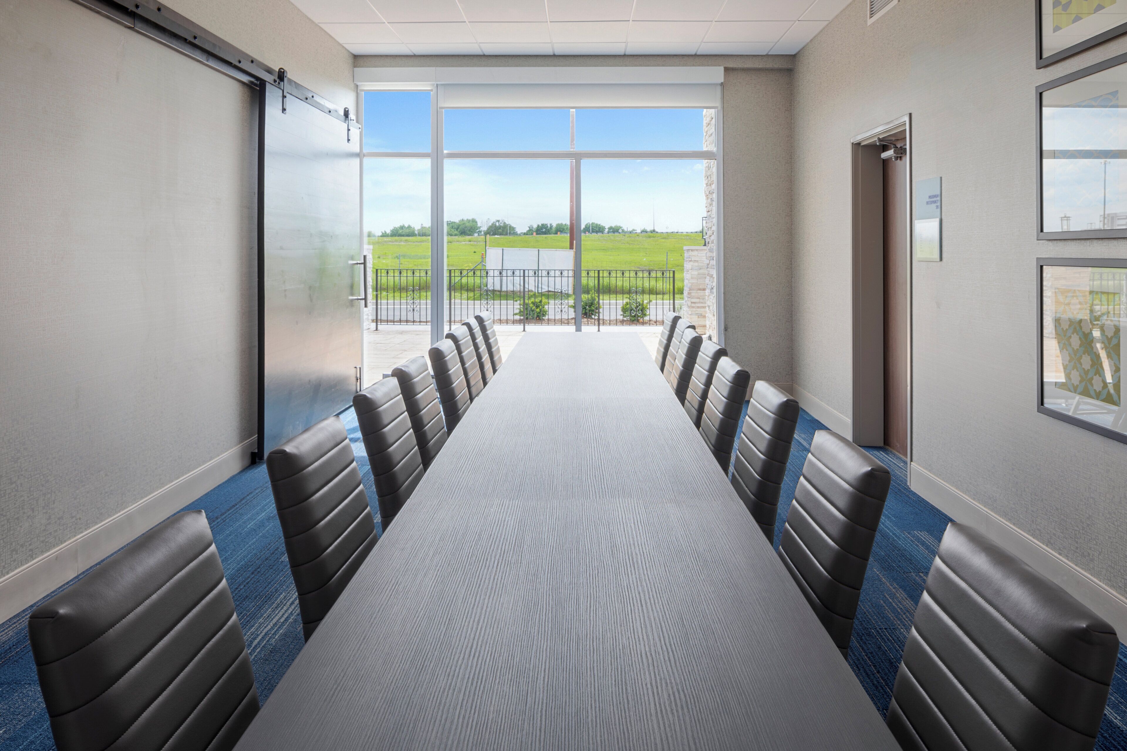 A conference room with a long table and chairs, large windows with a view of a green field.