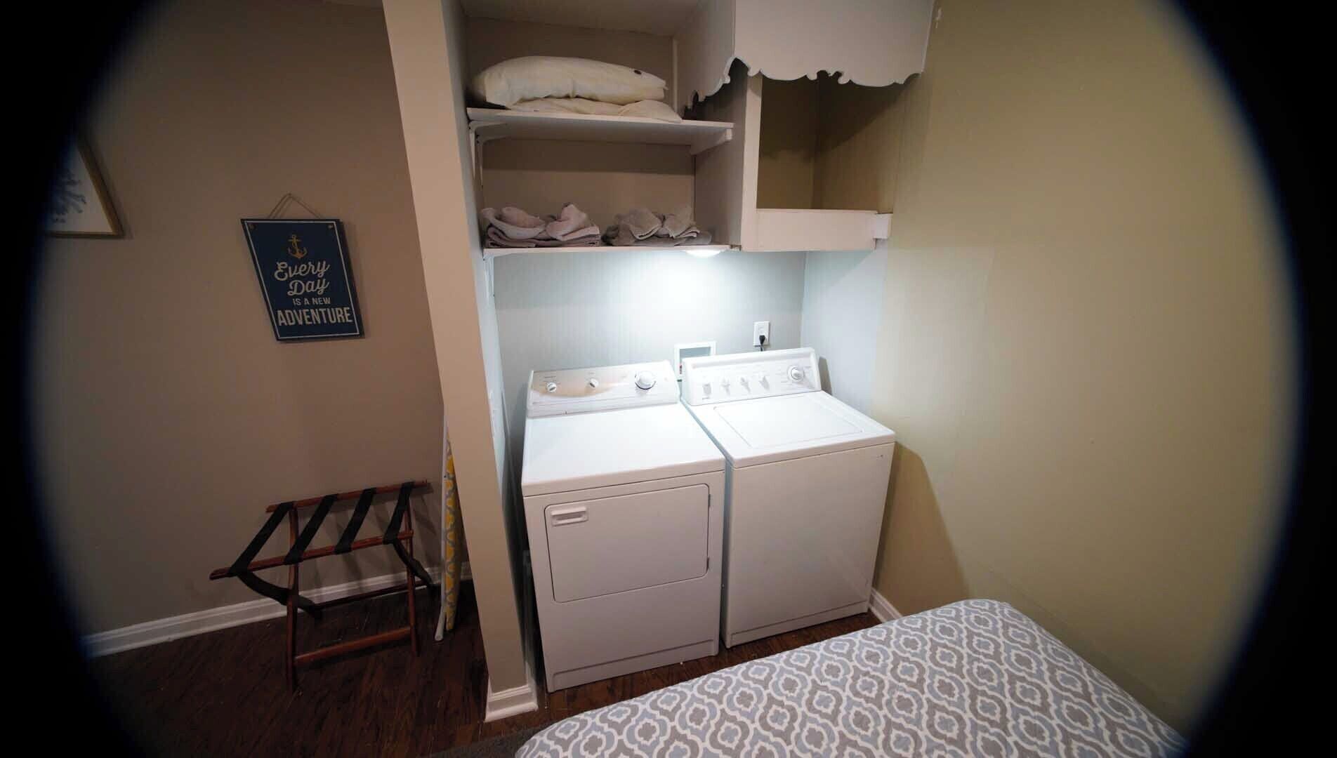 A compact laundry room with a washing machine and dryer, a small shelf with folded towels, and a motivational quote on the wall.